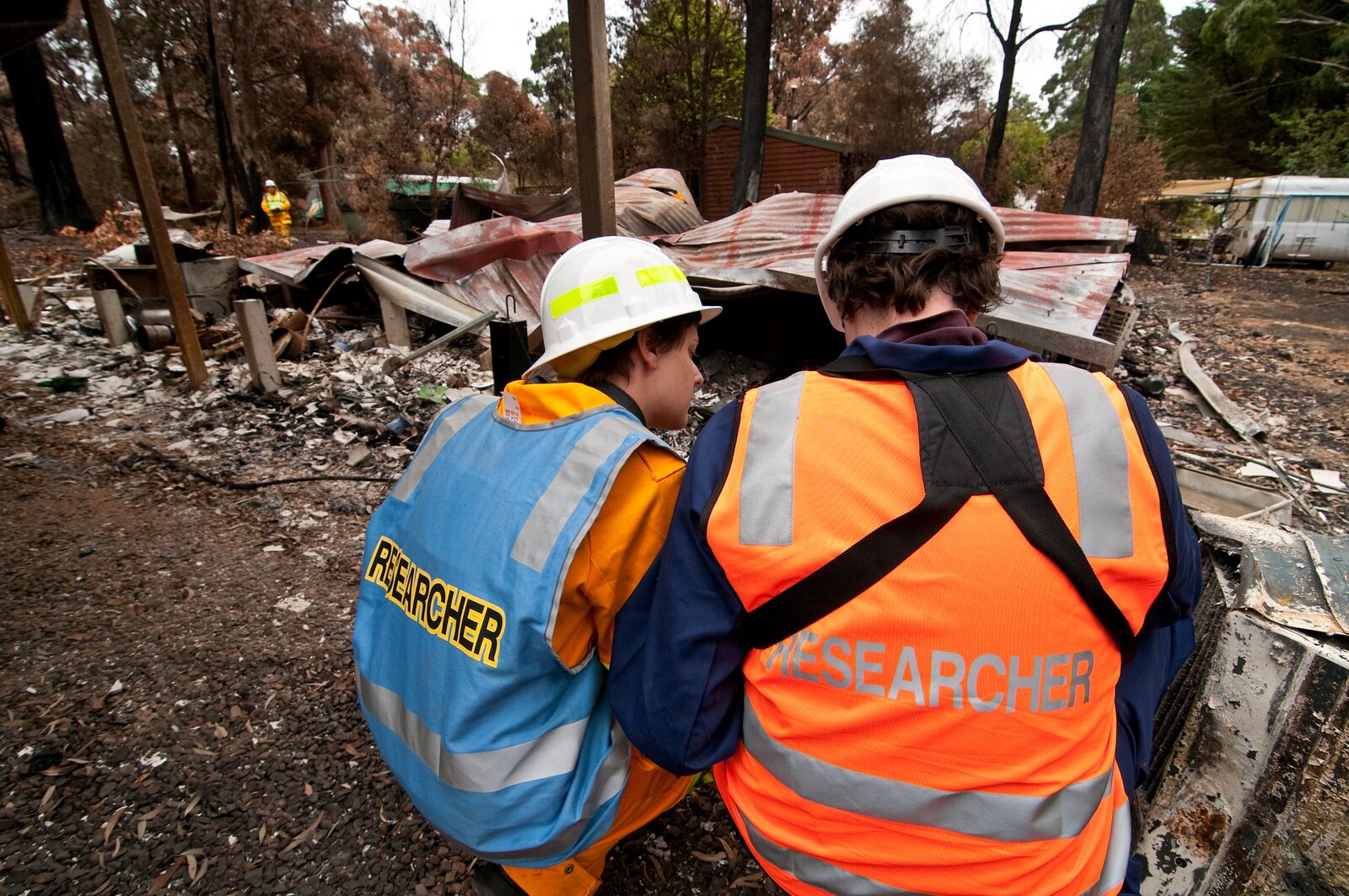 Two researchers in hard hats and high-visibility vests marked RESEARCHER crouch side by side, seen from behind, at a property destroyed by bushfire. Collapsed corrugated iron and ash cover the ground in front of them among burnt trees, with a caravan to the right and a firefighter in yellow in the background.