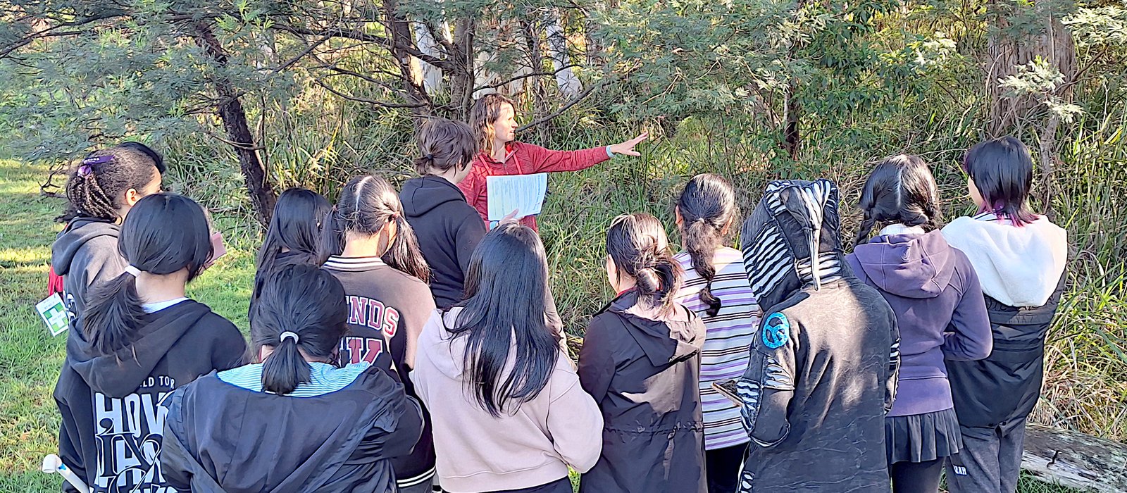 A group of about a dozen Year 11 students in jackets and hoodies, seen from behind, standing in long grass and facing a line of wattle and eucalypt scrub. In front of them Dr Martyna Kotowska, in a red top and holding a printed sheet, points into the understorey.
